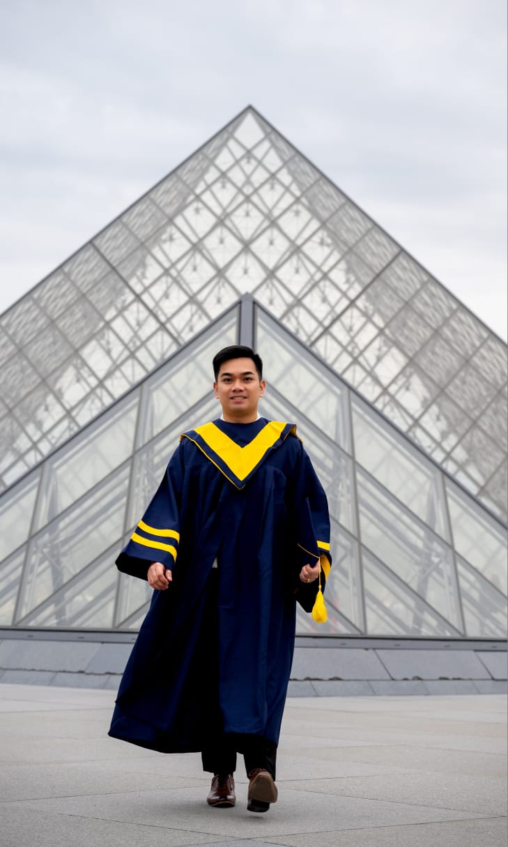 A graduate in cap and gown walks in front of the Louvre Pyramid in Paris