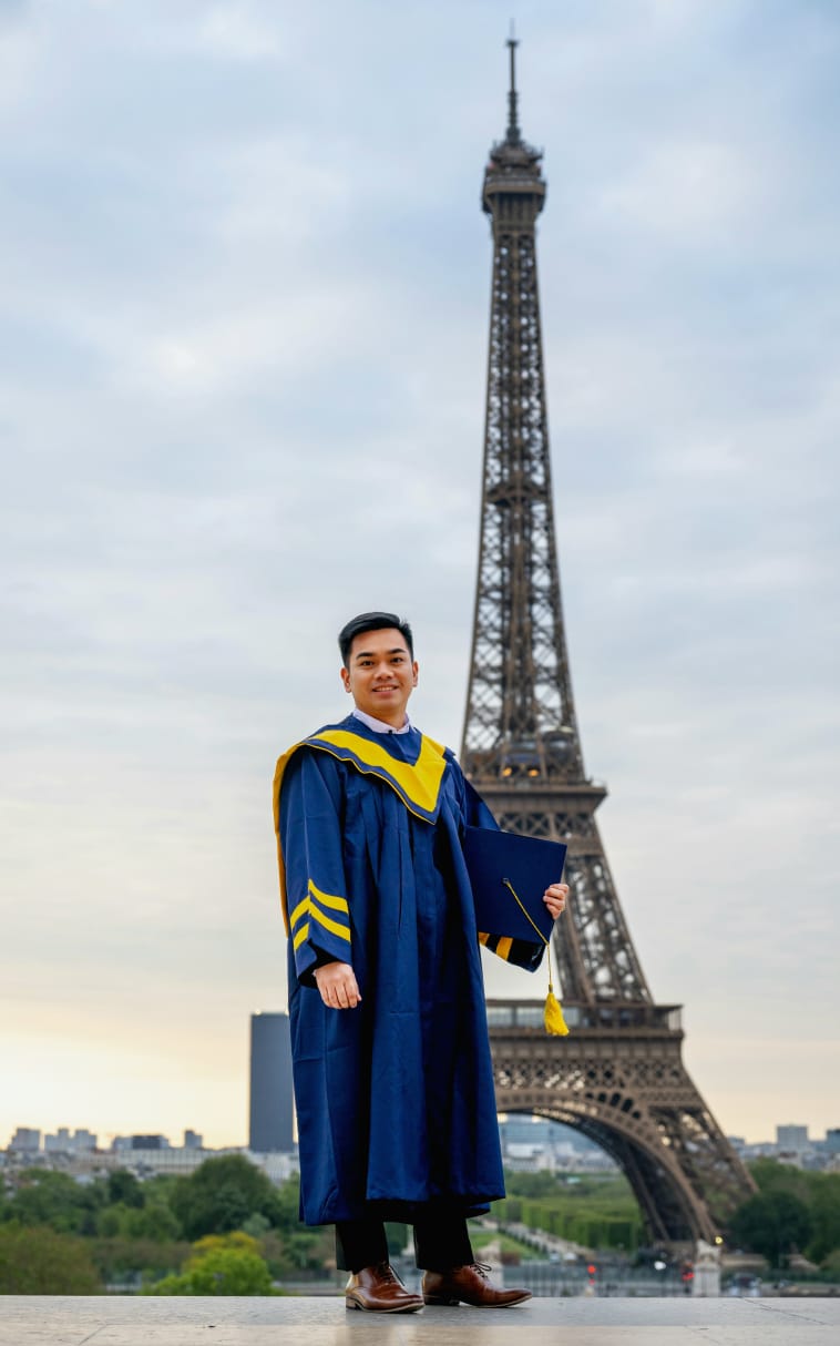 A graduate in cap and gown poses in front of the Eiffel Tower