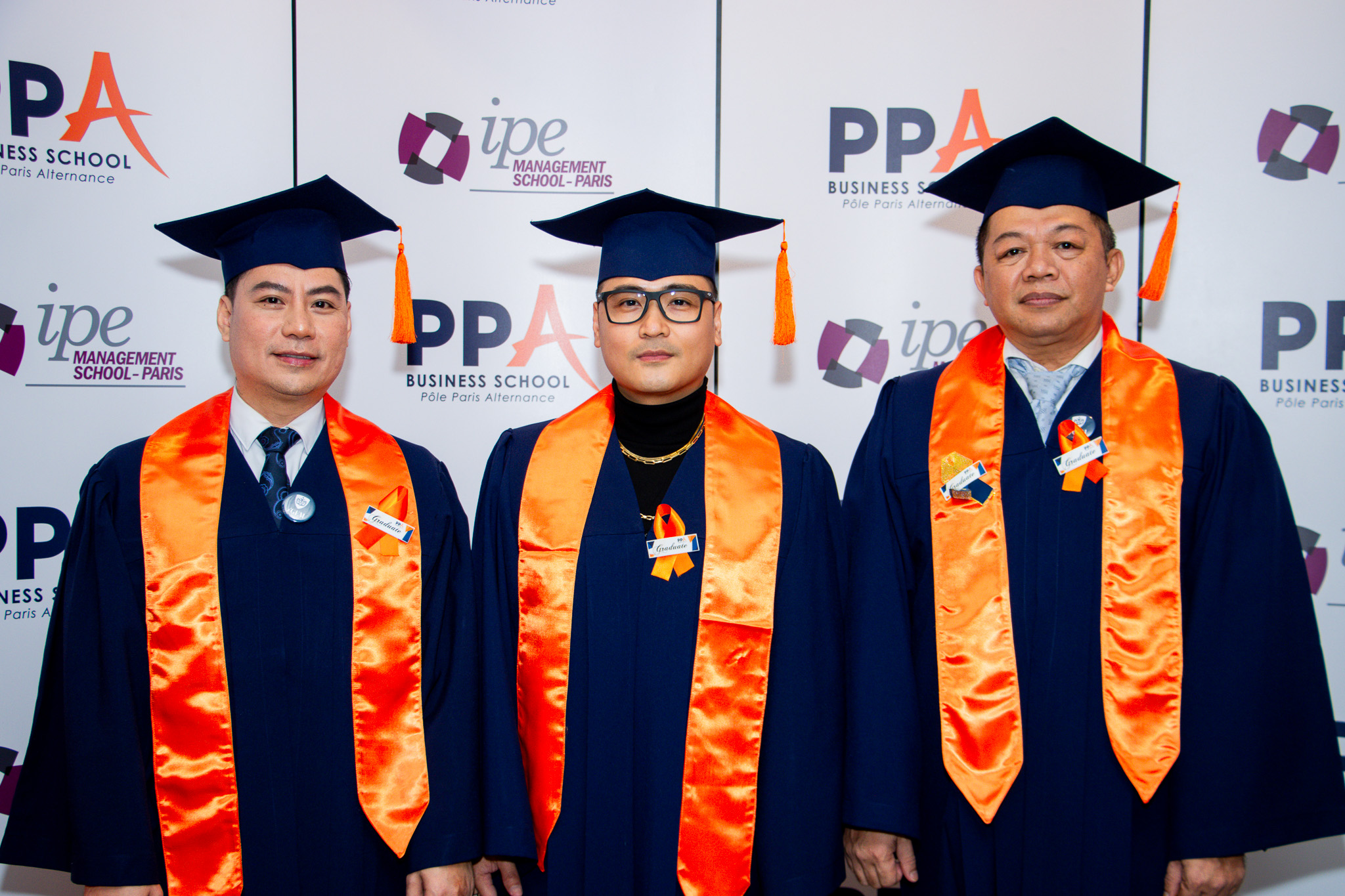 Three PPA Business School graduates in caps and gowns at the PPA-IPE step-and-repeat backdrop