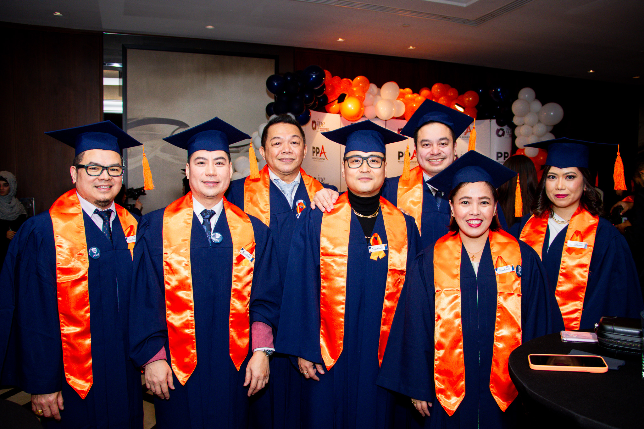 A group of PPA Business School graduates in navy gowns and orange sashes at the graduation reception